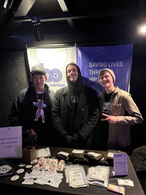 3 people stand together behind a table of overdose prevention materials and in front of blue and white posters reading "beatsoverdose" and "Saving Lives through Hip-hop"; 